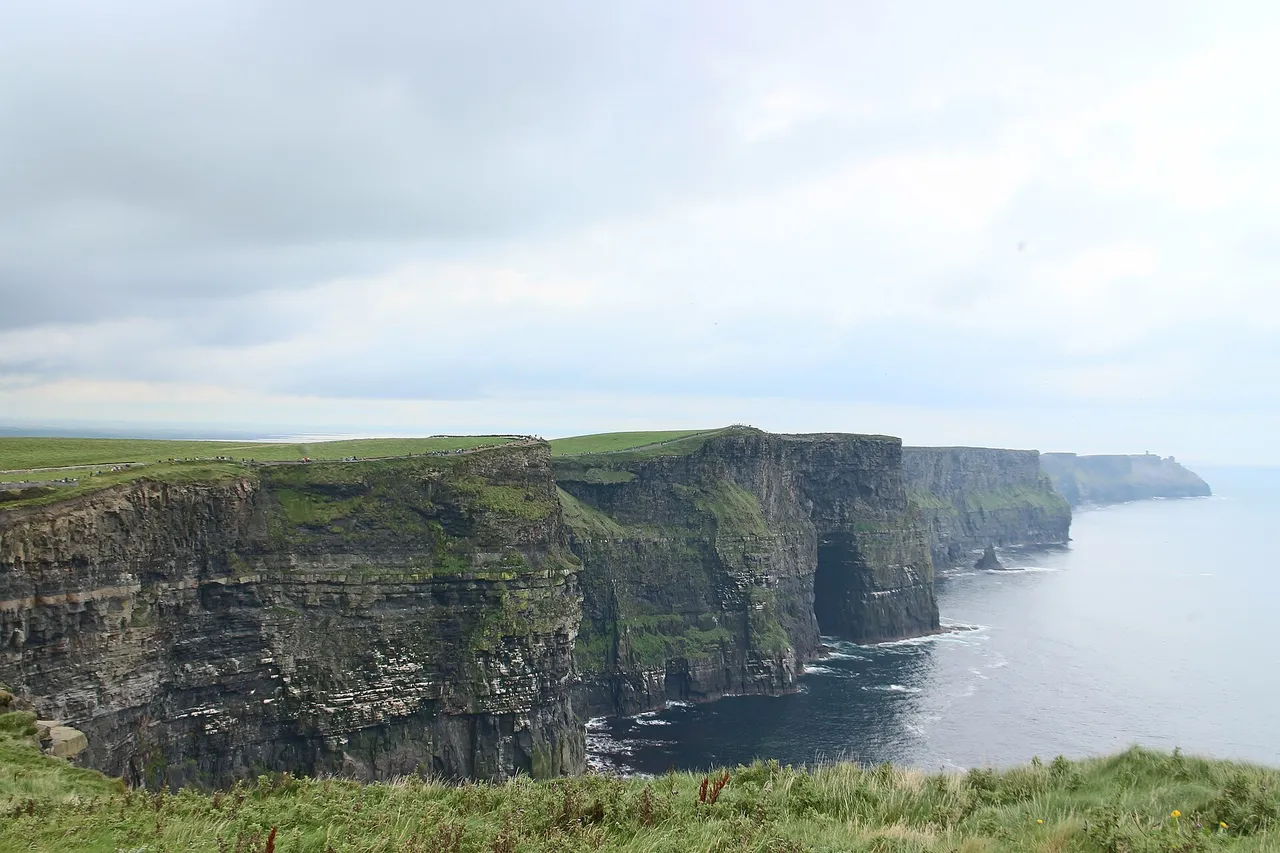 The dark, sheer Cliffs of Moher run along the Atlantic coast under a grey sky, with green grass on top and the sea far below