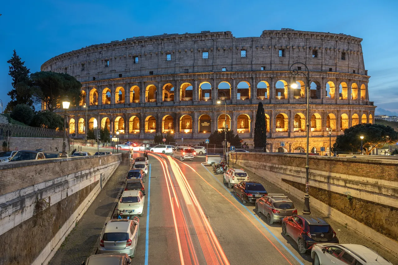 The Colosseum lit up at dusk, above a road with the light trails of passing cars