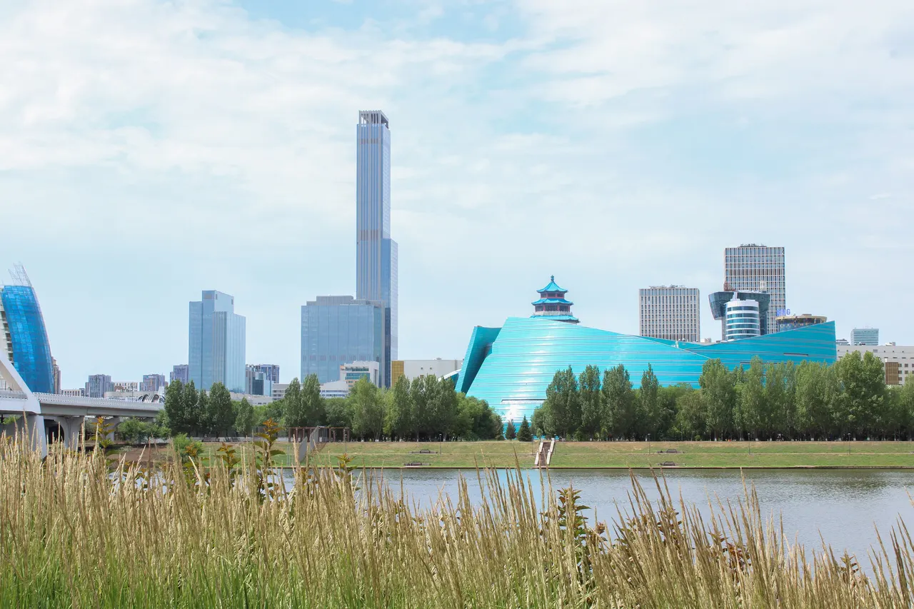 Glass towers and a curved turquoise building rise beyond the Esil River and tall grass in Astana, under a pale summer sky