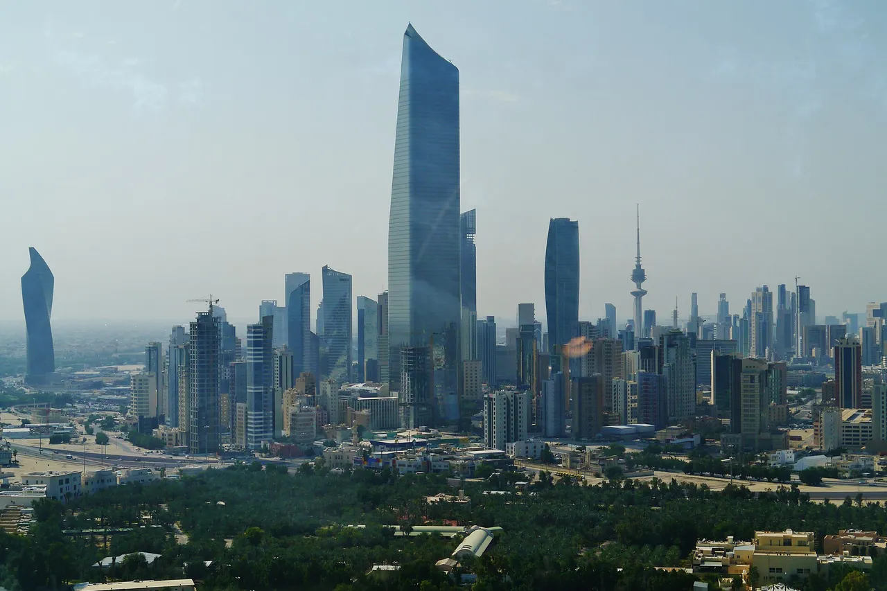 Glass skyscrapers of Kuwait City, led by the tall curved al-Hamra Tower, rise behind a belt of green trees