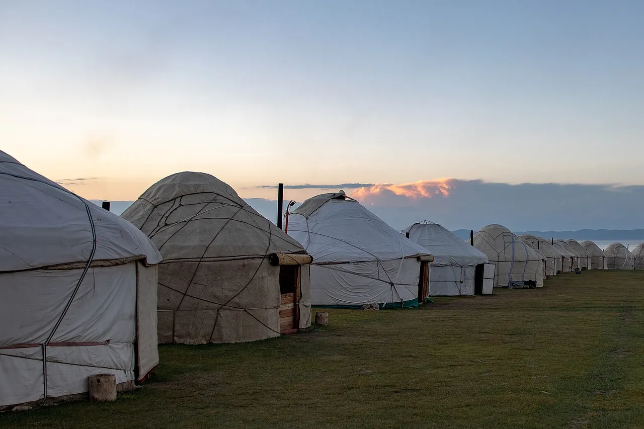 A long row of white felt yurts on green grassland at Song-Kul lake, with low mountains and a pale evening sky behind them