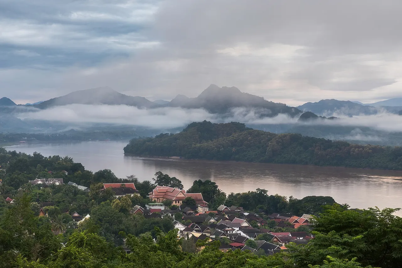 Low morning clouds hang over forested mountains and the brown Mekong River, with the tiled roofs of Luang Prabang among the trees in front