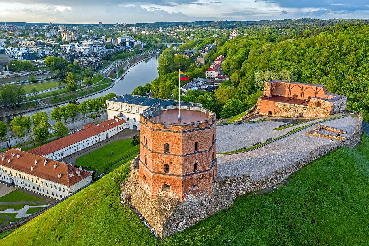 Gediminas Tower, a red-brick octagonal castle tower flying the Lithuanian flag, on a green hill above Vilnius, with the Neris river and the city behind