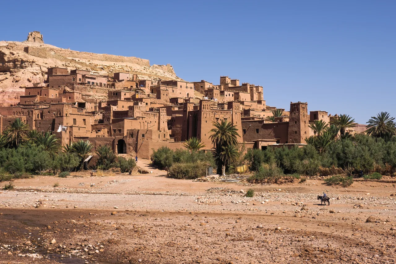 The mud-brick ksar of Ait Benhaddou climbing a hillside above palm trees and a dry riverbed