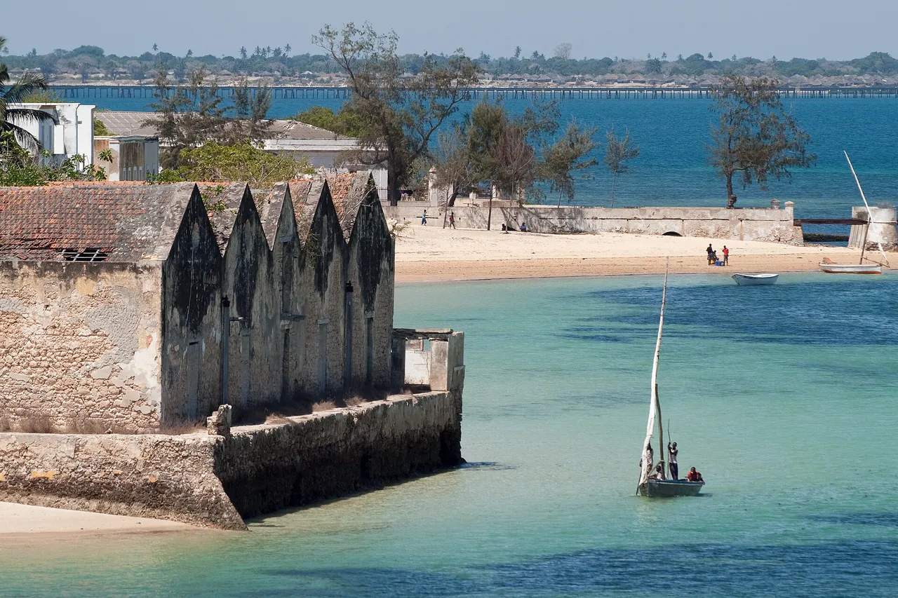 An old stone warehouse with a row of pointed gables stands on the shore of Mozambique Island, with a sailing dhow on turquoise water and the long bridge to the mainland behind