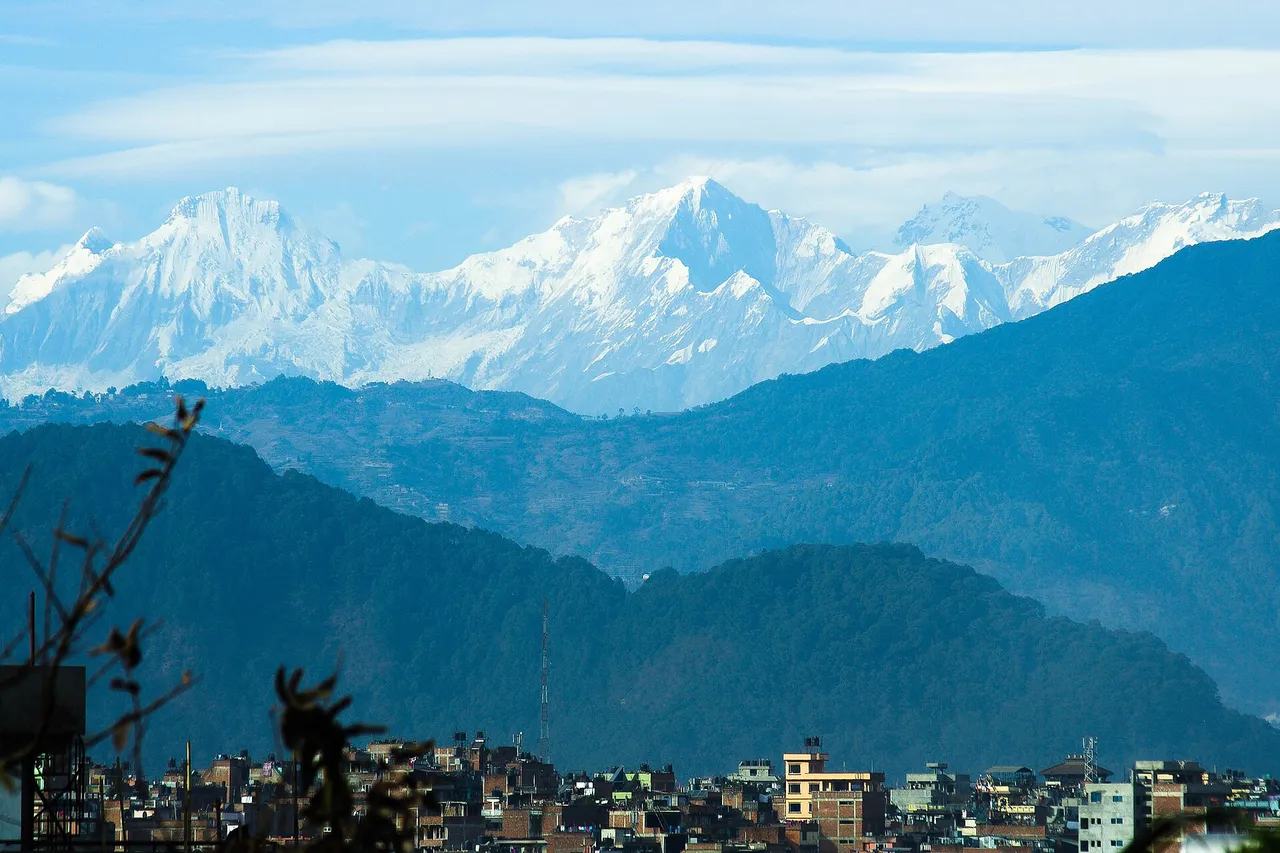 The snow-covered peaks of the Ganesh Himal rise above layers of blue-green hills, with the rooftops of Lalitpur in the Kathmandu valley in front