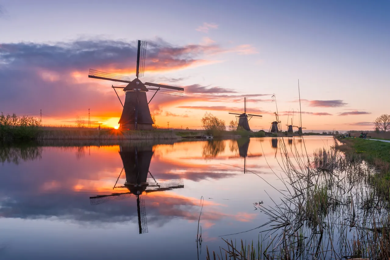A traditional windmill reflected in a still canal at sunrise, with a row of smaller windmills along the water behind it