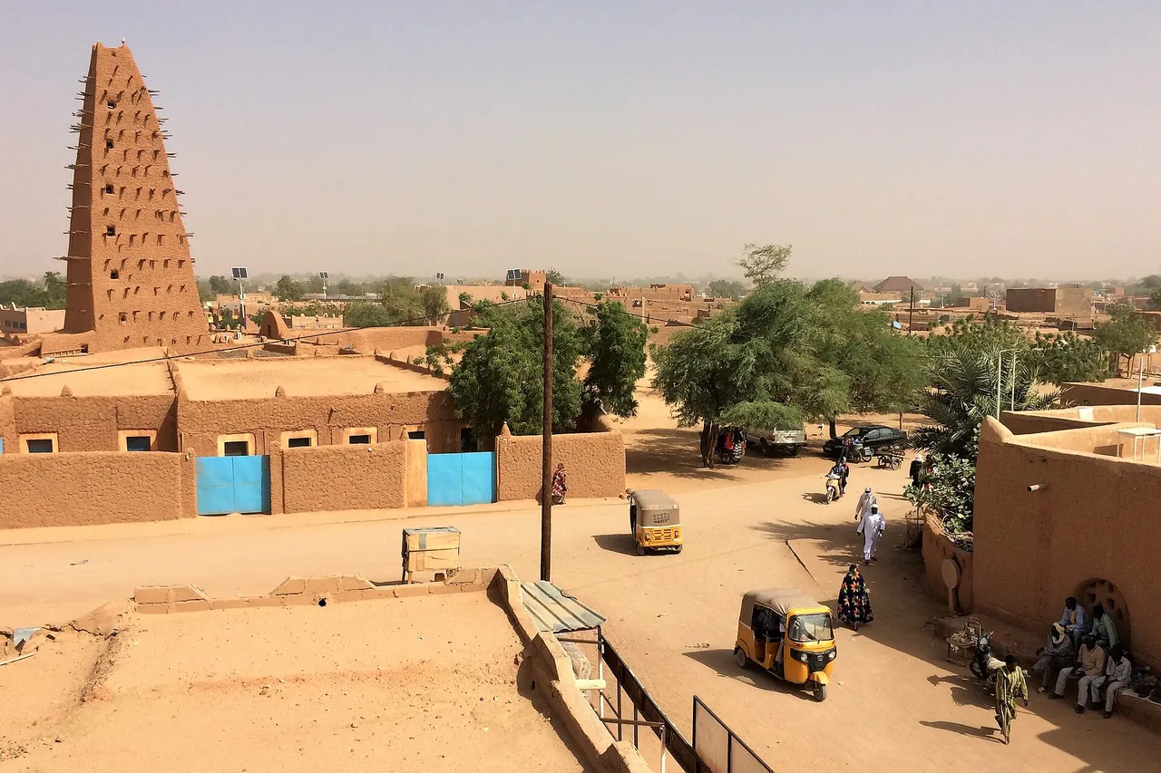 The tall mud-brick minaret of the Grand Mosque of Agadez rises above flat-roofed mud houses and a sandy street with motorized rickshaws