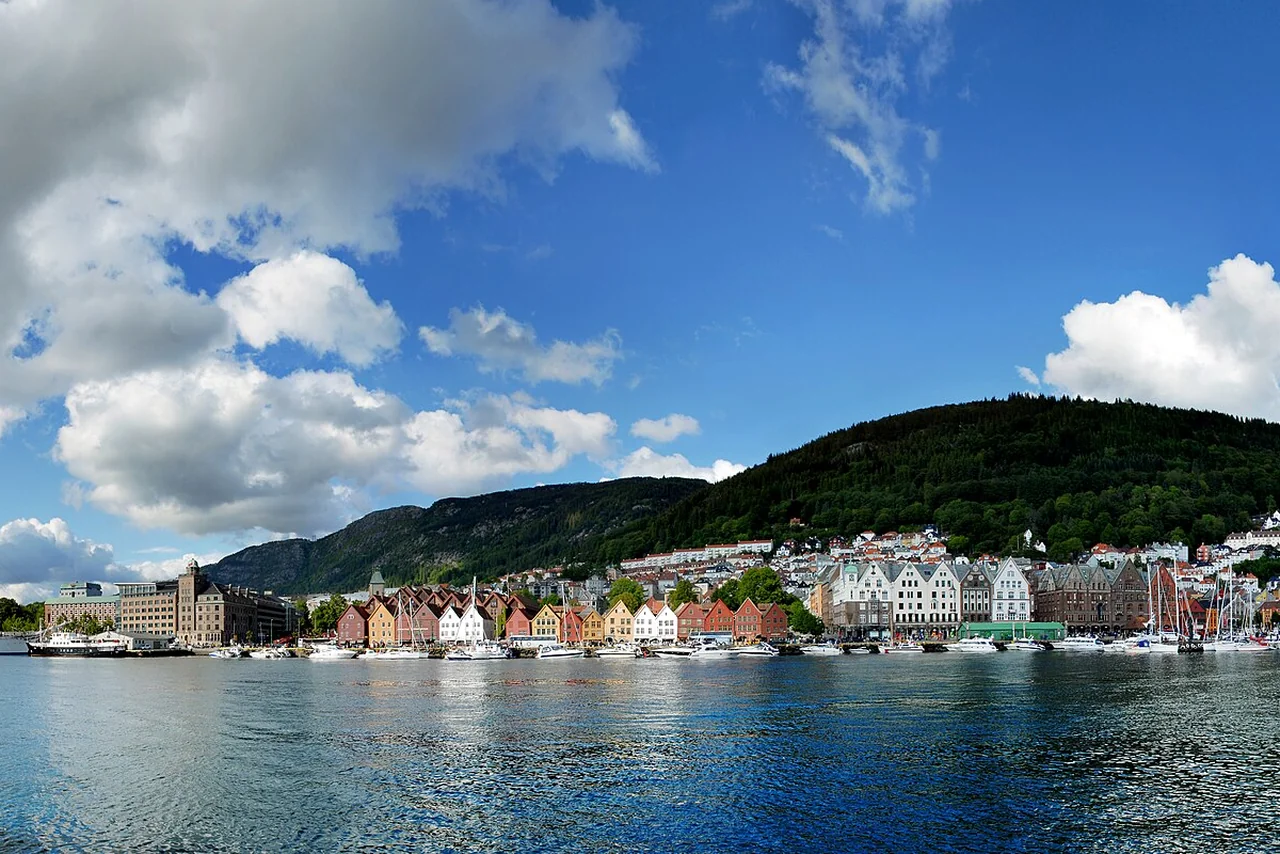 Colorful wooden buildings along Bergen's Bryggen waterfront, below a green hillside and reflected in the harbor