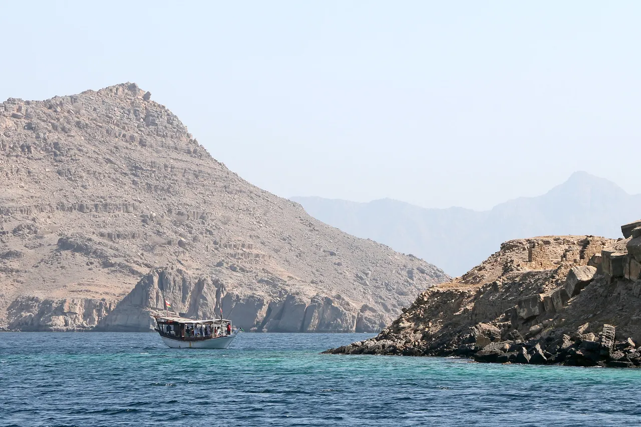 A wooden dhow full of tourists sails between bare rocky mountains in a turquoise inlet of the Musandam peninsula