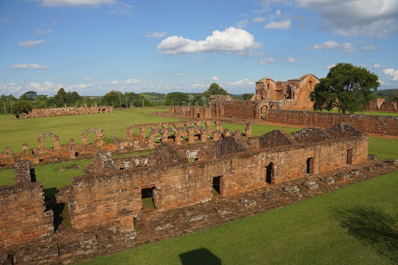 Red sandstone walls and arches of a ruined Jesuit mission stand on a green lawn, with the remains of a large church behind under a blue sky