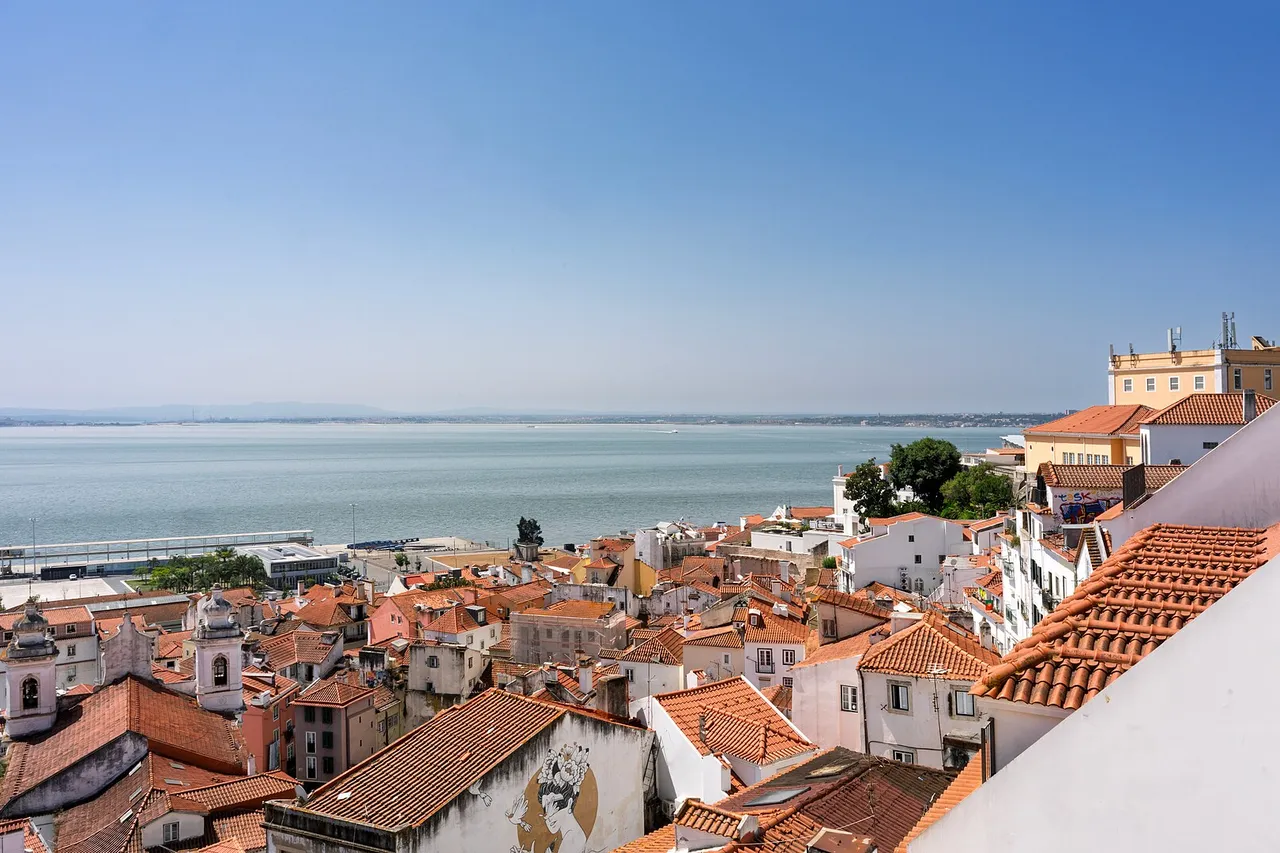 Terracotta rooftops and church towers of Lisbon's Alfama district slope down toward the wide Tagus River under a clear blue sky