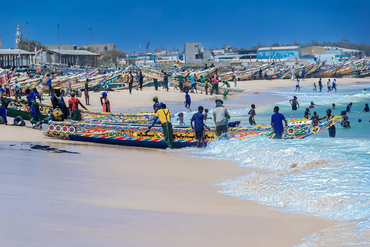 Fishermen push brightly painted wooden pirogues out of the surf onto a crowded sandy beach at Kayar, with more boats lined up behind