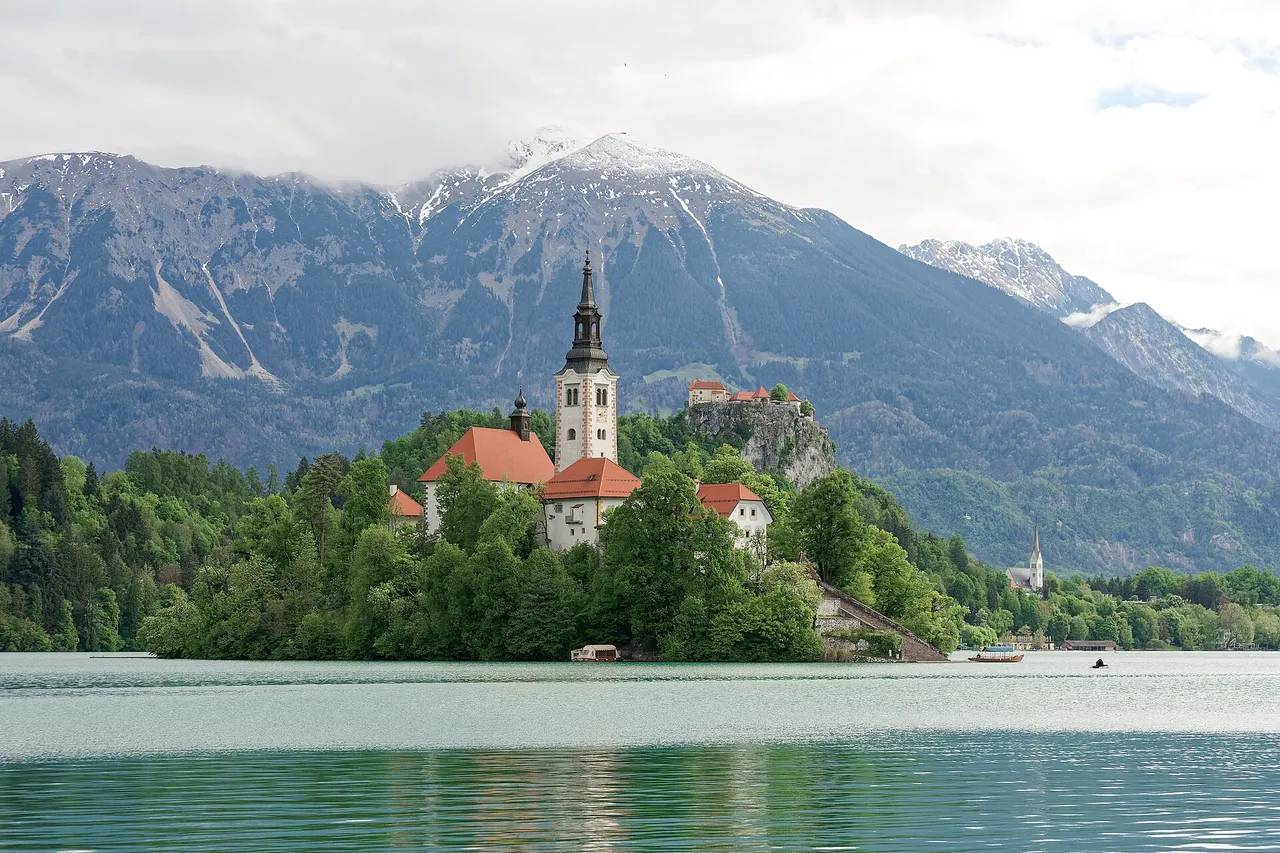 Bled Island with its white church tower in Lake Bled, Bled Castle on a cliff behind it and snowy Alpine peaks in the background