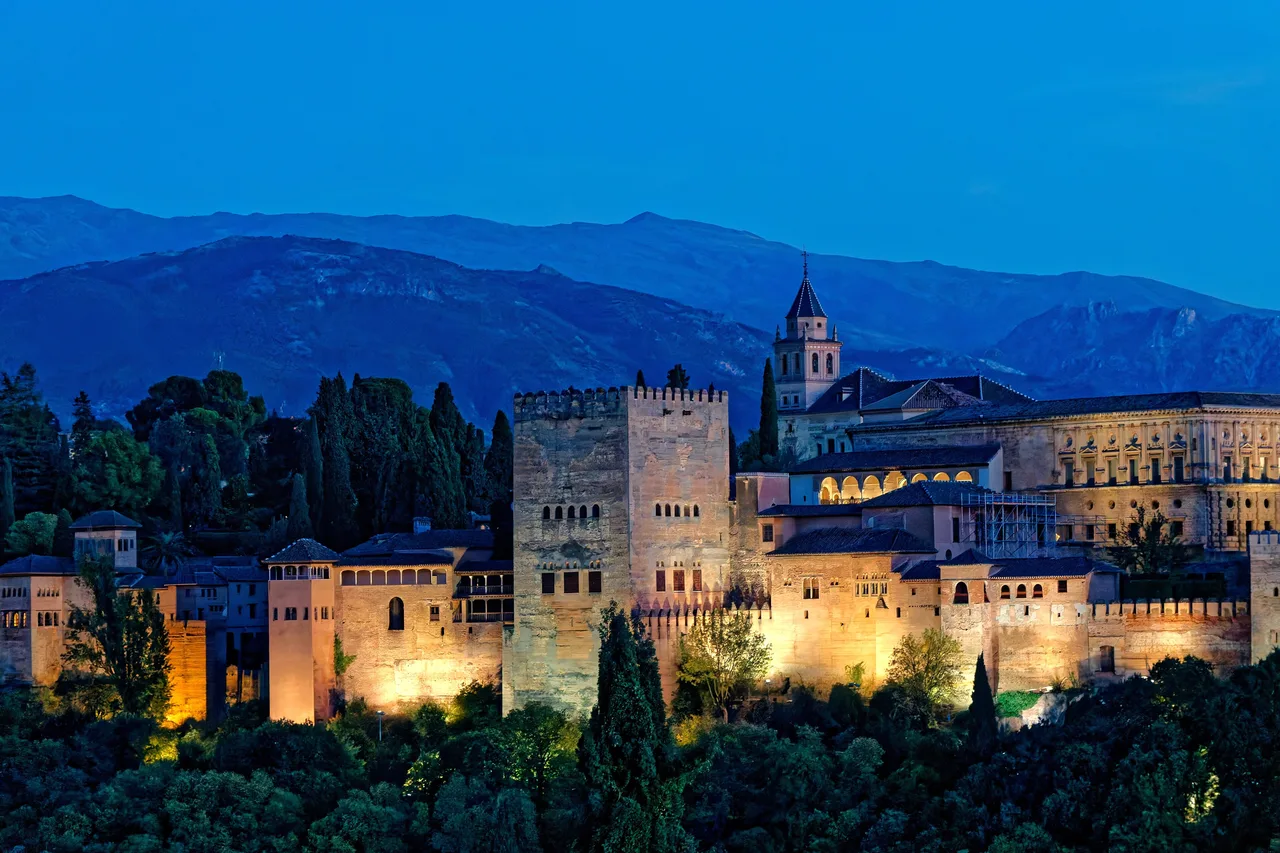 The Alhambra lit up at dusk on its wooded hill in Granada, with the Sierra Nevada behind
