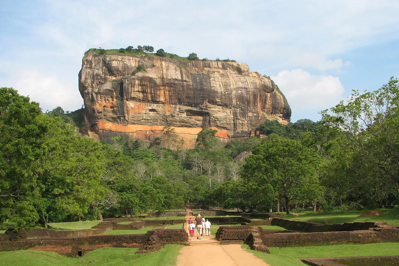 A huge flat-topped orange rock rising above green forest, with a path through the brick ruins of old royal gardens leading toward it