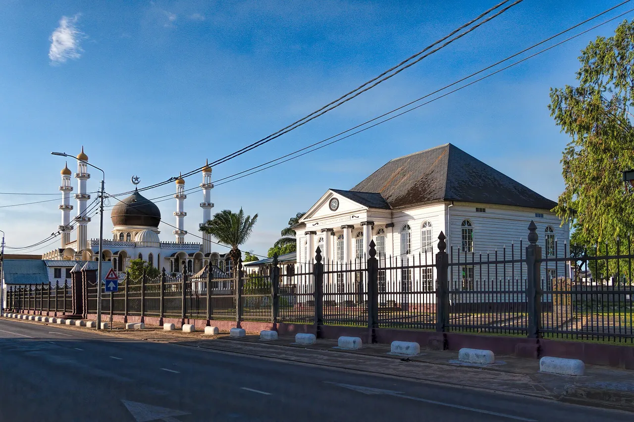 The white wooden Neveh Shalom synagogue next to a mosque with a dome and four minarets on Keizerstraat in Paramaribo, under a blue sky