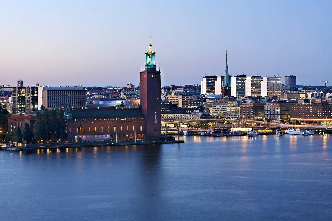 Stockholm City Hall with its tall brick tower on the water's edge at dusk, with the city lights reflected in the bay