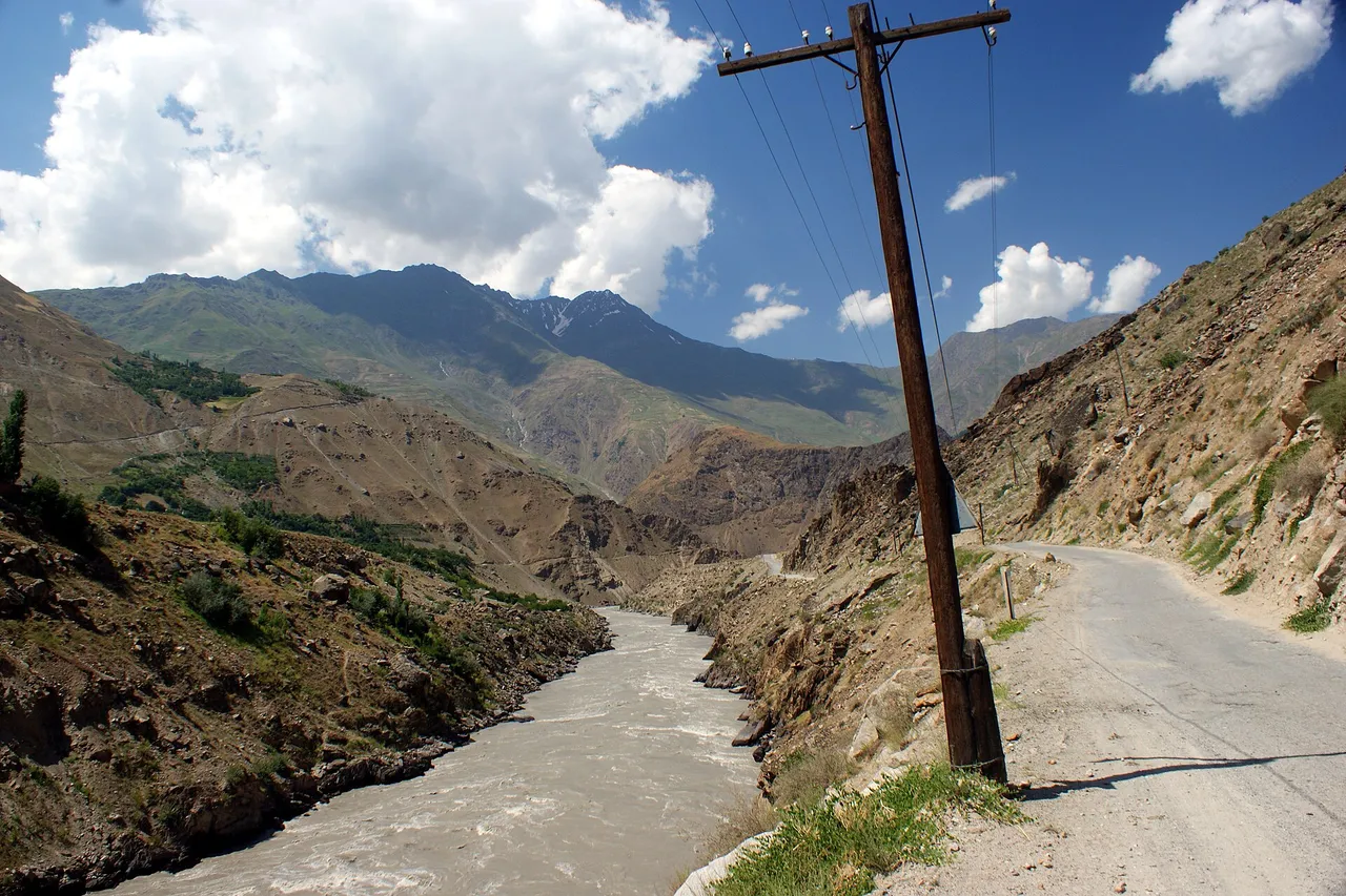 A dirt road and a wooden power pole run along the fast Panj River through a steep mountain valley, with Afghanistan on the far bank