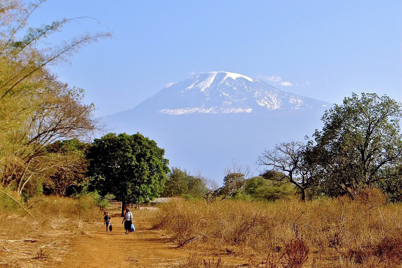 The snow-capped summit of Mount Kilimanjaro rises above dry grass and green trees, with two people walking along a dirt path in the foreground