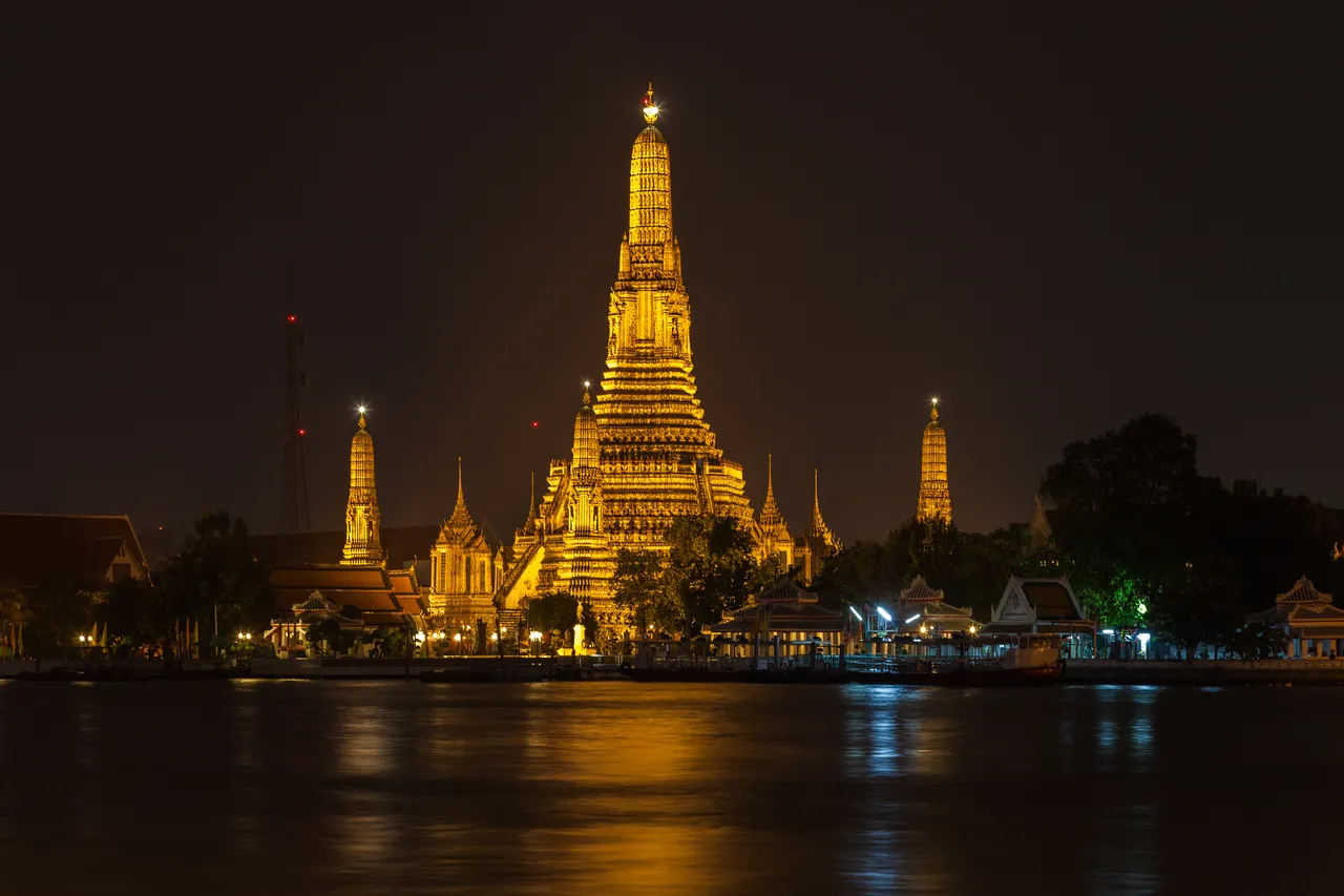 The tall central spire of Wat Arun, lit gold at night, with smaller spires around it, reflected in the dark Chao Phraya River