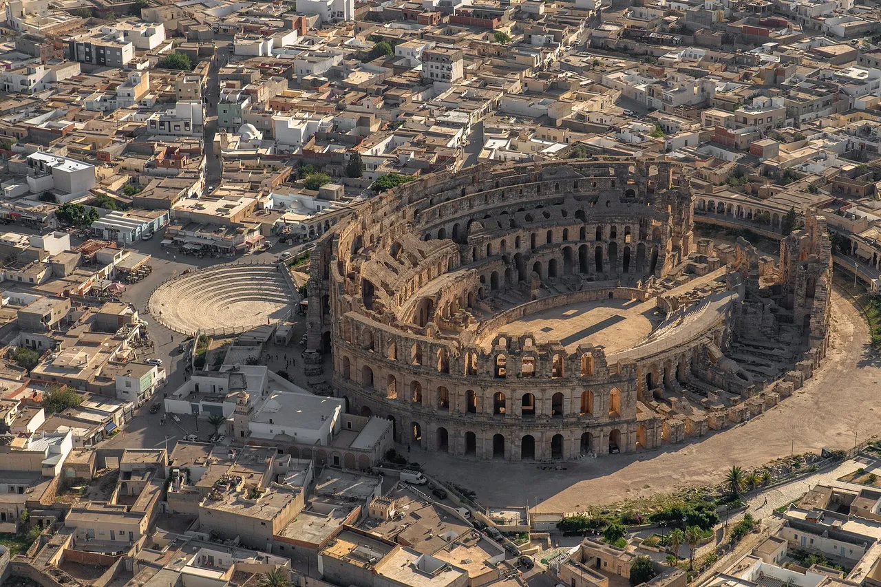 Aerial view of the Roman amphitheatre of El Jem, a huge oval of stone arches, surrounded by the low sand-colored houses of the town