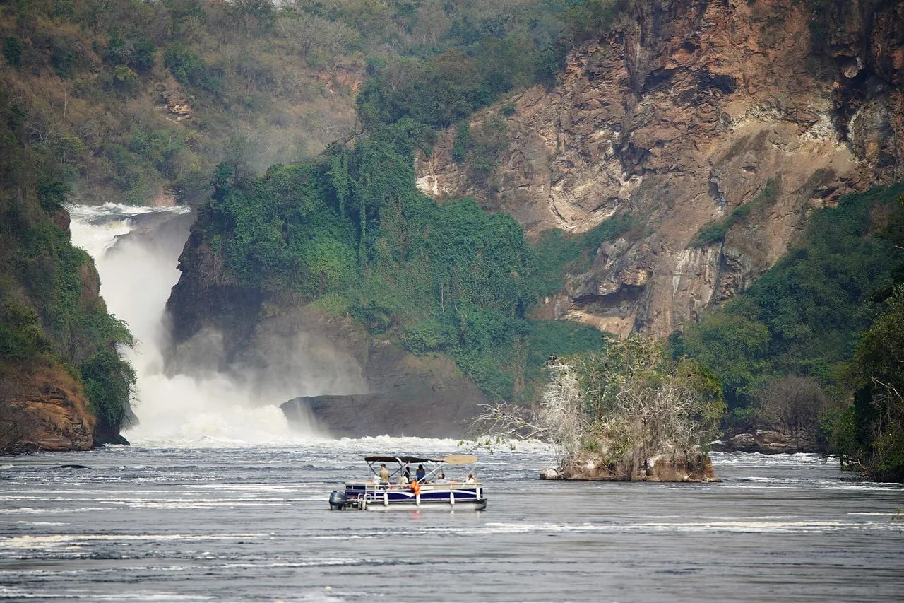 The Nile pours through a narrow gap in a rocky, forested gorge at Murchison Falls, with a tourist boat on the river below