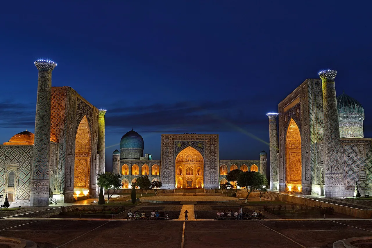 The Registan square in Samarkand at dusk, with three lit madrasas with tiled portals, minarets and a turquoise dome
