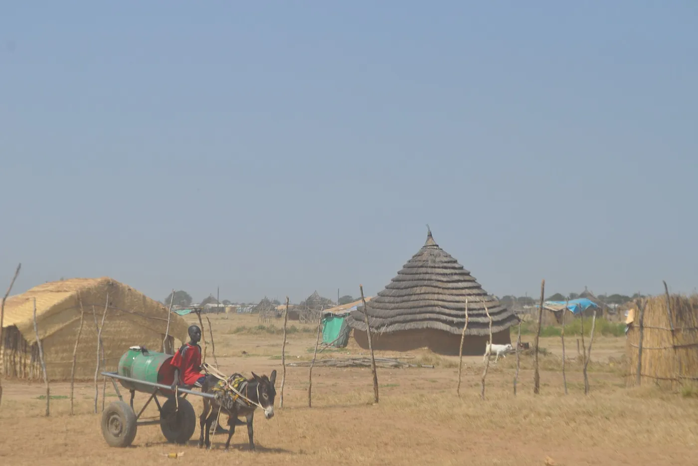 Round thatched huts in Abyei town, with a boy driving a donkey cart that carries a green water tank