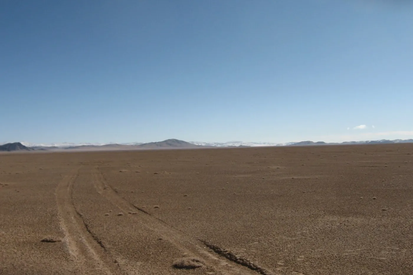 A flat, empty plain of brown gravel with tire tracks, under a blue sky, with low mountains on the horizon