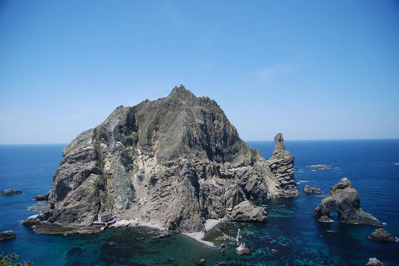 A steep, rocky islet rising from clear blue water, with a small house at its foot and rocks around it