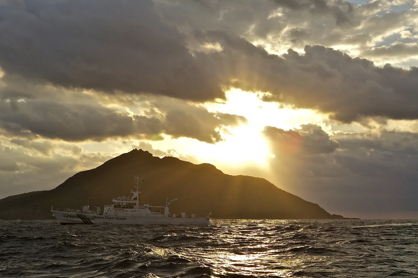 A Japan Coast Guard patrol ship passing the dark peak of Uotsuri island under clouds