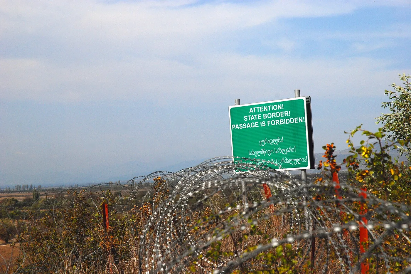 A green sign reading 'Attention! State border! Passage is forbidden!' behind coils of barbed wire in a field